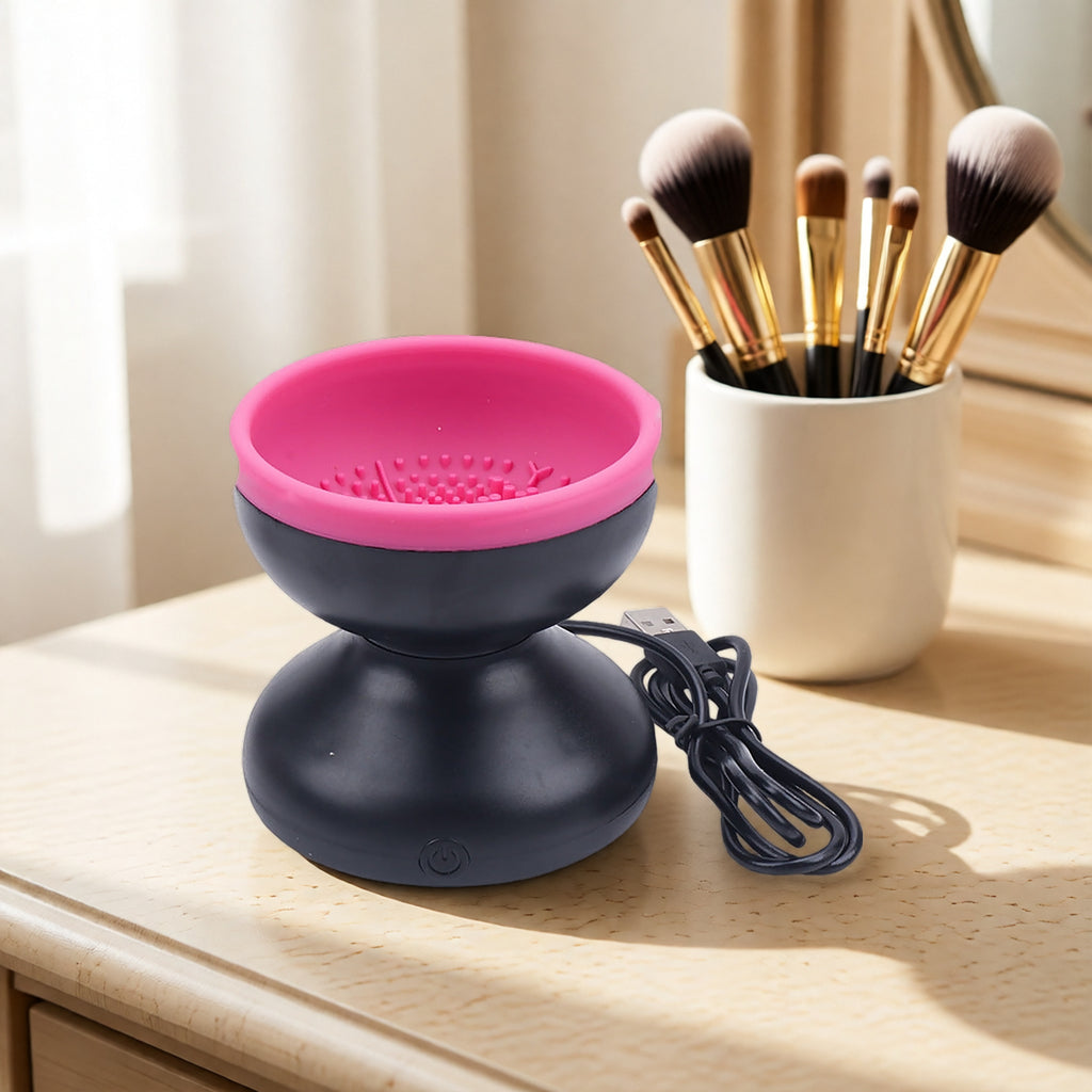 Pink and black electric makeup brush cleaner with USB cord on wooden vanity, next to white brush holder.