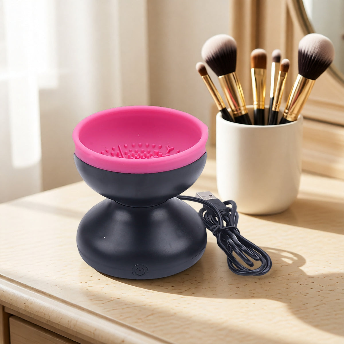 Pink and black electric makeup brush cleaner with USB cord on wooden vanity, next to white brush holder.